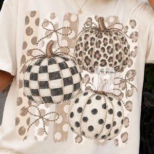 Könnte beinhalten: Ein cremefarbenes Kurzarm-T-Shirt mit einem Grafikdruck von drei Kürbissen. Ein Kürbis hat ein schwarz-weißes Karomuster, einer ein braunes Leopardenmuster und einer schwarze Punkte. Der Hintergrund zeigt beigefarbene Punkte.