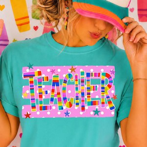 May include: A teal t-shirt featuring the word TEACHER in colorful, multi-striped block letters centered on a pink rectangular background with white polka dots and small star accents. The shirt is worn with a pink and teal baseball cap.