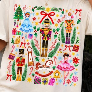Könnte beinhalten: Ein cremefarbenes T-Shirt mit einem bunten Grafikdruck zum Thema Weihnachten. Das Design zeigt Nussknacker, eine Ballerina, einen Maussoldaten, ein Schaukelpferd, eine Standuhr, verpackte Geschenke, Zuckerstangen und Tannenzweige.