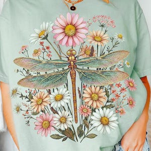 Pode incluir: Camiseta verde claro com um desenho de libélula e flores coloridas. A libélula tem asas detalhadas e é cercada por margaridas e outras flores em tons de rosa, amarelo e branco. O texto "DAVINCIPRINTSHOP" e "ORIGINAL DESIGNER" estão no topo.