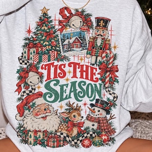 May include: A light gray crewneck sweatshirt featuring a colorful Christmas-themed graphic print. The design includes a nutcracker, Santa Claus, a reindeer, a snowman, a decorated tree, gifts, and the text 'TIS THE SEASON' in red and green.