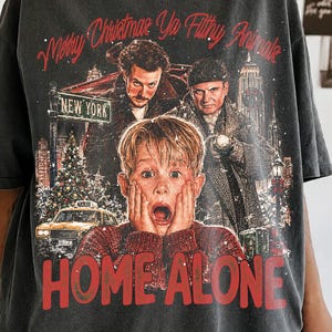 May include: A charcoal grey t-shirt featuring a graphic print from the movie Home Alone. The design includes the text Merry Christmas Ya Filthy Animals, a central image of a boy with hands on his face, two men, a taxi, and the text HOME ALONE.