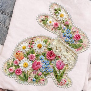 May include: A white t-shirt featuring an embroidered bunny design. The bunny is filled with colorful flowers, including daisies, roses, and tulips, and is outlined with pearls. The text "DAVINCIPRINTSHOP" and "ORIGINAL DESIGNER" are visible.