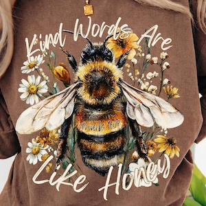 Könnte beinhalten: Braunes Sweatshirt mit einer großen Illustration einer Biene, umgeben von Blumen. Der Text "Kind Words Are Like Honey" und "Proverbs 16:24" ist auf dem Shirt aufgedruckt. Das Sweatshirt hat einen Rundhalsausschnitt.