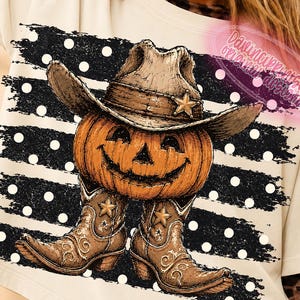 K&ouml;nnte beinhalten: Ein cremefarbenes T-Shirt mit einer Grafik eines orangefarbenen K&uuml;rbisses, der einen braunen Cowboyhut und braune Cowboystiefel tr&auml;gt. Das Design ist vor einem Hintergrund aus schwarzen horizontalen Pinselstrichen mit wei&szlig;en Punkten platziert.