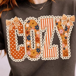 May include: A dark brown crewneck sweatshirt featuring the word COZY in large, appliqued letters. Each letter has a unique pattern including white polka dots, orange floral prints, and orange stripes, all bordered by cream-colored lace trim.
