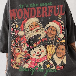 K&ouml;nnte beinhalten: Ein anthrazitfarbenes Oversize-T-Shirt mit einem Grafikdruck und dem Text "it's the most WONDERFUL time of the year". Das Design zeigt Illustrationen vom Weihnachtsmann, einem Rentier, einem Schneemann und Buddy dem Elf.