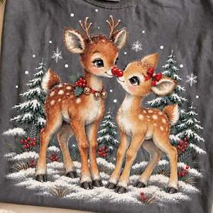 Peut inclure: Un t-shirt à col rond gris anthracite orné d'une illustration graphique de deux faons bruns dans une forêt enneigée. Un faon porte un collier avec des clochettes dorées et des baies rouges, l'autre a un nœud rouge à l'oreille. Flocons et sapins.