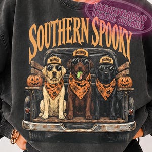 May include: A charcoal gray crewneck sweatshirt featuring a graphic of three dogs sitting in the bed of a vintage truck. The dogs wear Halloween-themed bandanas, sunglasses, and baseball caps. The text reads SOUTHERN SPOOKY in orange letters.