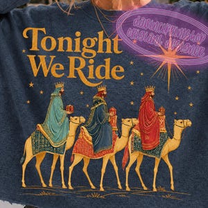 以下が含まれることがあります： ゴールドの文字で「Tonight We Ride」と書かれたダークブルーの長袖スウェットシャツ。王冠とローブを身にまとった3人の人物がそれぞれラクダに乗っている様子が描かれており、その上には星のデザインと小さな金色の星が散りばめられています。