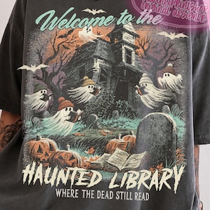 May include: A charcoal grey t-shirt featuring a graphic of a haunted house, ghosts wearing beanies and reading books, pumpkins, and a tombstone. The text reads: Welcome to the HAUNTED LIBRARY WHERE THE DEAD STILL READ. DAVINCIPRINTSHOP is printed at the top.