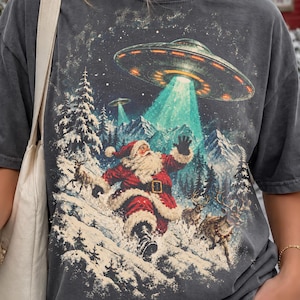 May include: A charcoal grey t-shirt featuring a graphic print of Santa Claus in a snowy landscape with reindeer, looking up at a large UFO emitting a bright green light beam. The design includes snowy mountains and pine trees in the background.