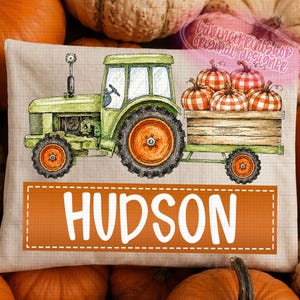Op de afbeelding: Een rechthoekig kussen met een groene tractor die een houten aanhanger vol oranje-wit geblokte pompoenen trekt. De naam HUDSON staat in grote witte letters in een oranje rechthoekig kader op een beige achtergrond met een raster.