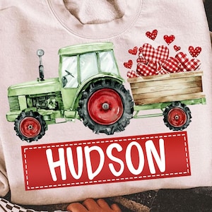 May include: A pale pink sweatshirt with a green tractor and red wheels, towing a wooden cart filled with red and white checked hearts. The name "HUDSON" is in white on a red rectangular banner.