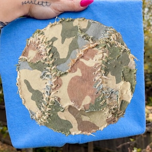 Può includere: Una maglietta azzurra con un grande motivo mimetico circolare. Il motivo mimetico presenta sfumature di verde, marrone e beige. Il testo "DAVINCIPRINTSHOP" e "ORIGINAL DESIGNER" sono visibili nella parte superiore dell'immagine.