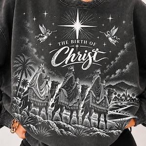 Peut inclure: Un sweat-shirt à col rond gris anthracite avec une impression graphique blanche représentant les trois rois mages à dos de chameau vers une ville lointaine. Le texte THE BIRTH OF CHRIST apparaît sous une étoile et deux anges.