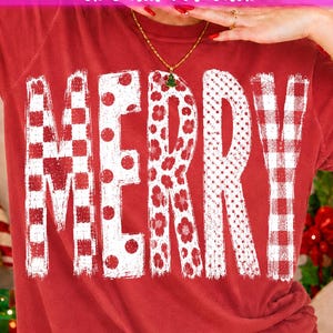 Peut inclure: Un t-shirt rouge à manches courtes avec le mot MERRY en grandes lettres blanches. Chaque lettre présente un motif distinct : damier rouge et blanc, pois rouges, motifs floraux rouges, petits points blancs et carreaux vichy rouges et blancs.