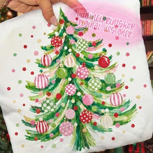 Puede incluir: Una sudadera blanca con un árbol de Navidad verde estilo acuarela, decorado con varios adornos estampados en rosa, rojo y verde. El diseño incluye puntos coloridos dispersos y el texto Davinciprintshop Original Designer.