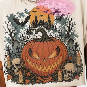 May include: A cream-coloured t-shirt featuring a graphic print of a large orange jack-o'-lantern, two human skulls, lit candles, and a spooky castle silhouette under a full moon with bats. The text DAVINCIPRINTSHOP appears at the top.
