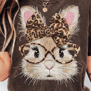 May include: Brown t-shirt featuring an embroidered bunny face with glasses and a leopard print bow. The bunny has pink inner ears and long eyelashes. The t-shirt is a dark brown color.