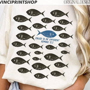 May include: Cream-colored t-shirt featuring a design of black fish swimming in various directions, with one blue fish standing out. The text "CALLED TO BE DIFFERENT ROMANS 12:2" is printed below the blue fish. The shirt has short sleeves.