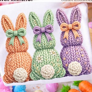 May include: A white fabric featuring three knitted Easter bunny designs in orange, green, and purple. Each bunny has a bow and a fluffy tail. The text "DAVINCIPRINTSHOP" and "ORIGINAL DESIGNER" are visible.