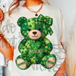May include: Cream-colored t-shirt featuring a green patchwork teddy bear design. The bear has a bow, shamrocks, and the words "Lucky", "Shamrock", and "Irish". The design is in shades of green and brown.