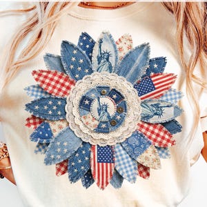 Pode incluir: Camiseta creme com um design patriótico de girassol. As pétalas são feitas de ganga, xadrez e tecidos florais em vermelho, branco e azul. O centro apresenta a Estátua da Liberdade. Os textos "DAVINCIPRINTSHOP" e "ORIGINAL DESIGNER" são visíveis.