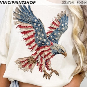 Pu&ograve; includere: T-shirt color crema con un design di aquila della bandiera americana invecchiata. Le ali dell'aquila sono spiegate, mostrando strisce rosse e bianche e toppe blu con stelle bianche. I testi "DAVINCIPRINTSHOP" e "ORIGINAL DESIGNER" sono visibili.