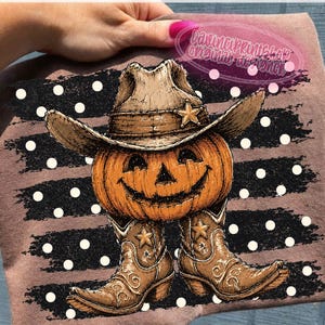 K&ouml;nnte beinhalten: Ein braunes T-Shirt mit einer Grafik eines orangefarbenen K&uuml;rbisses, der einen beigen Cowboyhut und braune Cowboystiefel tr&auml;gt. Das Motiv ist vor einem Hintergrund aus schwarzen horizontalen Pinselstrichen mit wei&szlig;en Punkten platziert.
