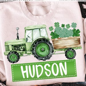 May include: A light pink sweatshirt featuring a green tractor pulling a wooden cart filled with shamrocks. The name "HUDSON" is displayed in white letters on a green rectangle. The text "DAVINCIPRINTSHOP" and "ORIGINAL DESIGNER" are at the top.