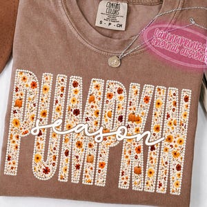 May include: A brown short-sleeved t-shirt featuring the word PUMPKIN in large block letters filled with a floral pattern of orange, yellow, and brown flowers and pumpkins, with the word season written in white cursive script across the center.