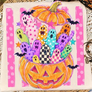 May include: A beige t-shirt featuring a graphic print of a large orange jack-o'-lantern filled with colorful, patterned ghosts. The design includes pink vertical stripes with white polka dots, small black bats, and various stars in the background.