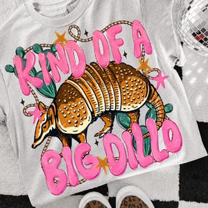 May include: A light gray t-shirt with a graphic of an armadillo and the text "KIND OF A BIG DILLO" in pink bubble letters. The armadillo is brown with yellow accents, surrounded by cactus and stars.
