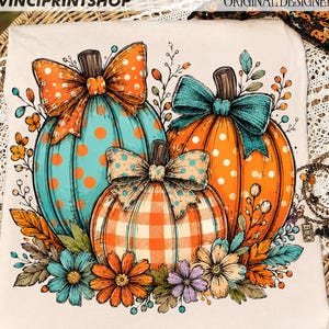 May include: A cream-colored t-shirt featuring a graphic of three pumpkins decorated with bows. The pumpkins are teal with orange dots, orange with white dots, and orange-and-white gingham. The design includes autumn flowers and leaves.