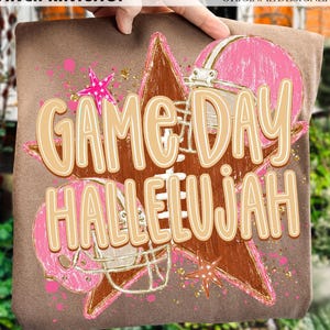 May include: A brown t-shirt featuring a graphic print of a large brown star, two pink football helmets, and pink star accents. The text "GAME DAY HALLELUJAH" is printed in large, light tan letters across the center of the graphic design.