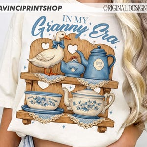 Puede incluir: Camiseta de color crema con una ilustraci&oacute;n caprichosa de un pato, teteras y tazas en una estanter&iacute;a de madera. La frase "IN MY Granny Era" est&aacute; impresa en azul sobre el dise&ntilde;o. El dise&ntilde;o incluye patrones florales azules y blancos.