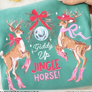 May include: Teal sweatshirt with a festive design of two deer in cowboy hats and boots. The text "Giddy Up Jingle Horse!" is below a disco ball and red bow. The words "DAVINCIPRINTSHOP" and "ORIGINAL DESIGNER" are at the bottom of the image.