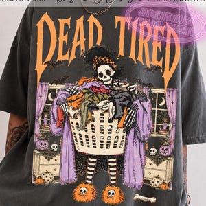 May include: A charcoal gray t-shirt featuring a graphic of a skeleton wearing a leopard print headband and fuzzy orange slippers, holding a laundry basket filled with clothes. The text DEAD TIRED is printed in large orange letters above the skeleton.