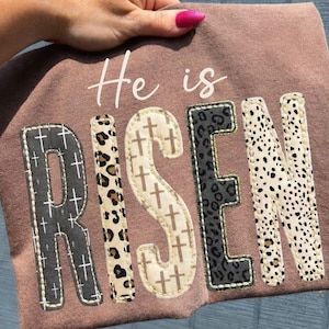 Puede incluir: Una camiseta de color malva con la frase "He is RISEN" en una fuente decorativa. Las letras de "RISEN" tienen patrones únicos, incluyendo cruces, estampado de leopardo y manchas de dálmata. La camiseta es sostenida por una mano.