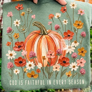 May include: A sage green sweatshirt featuring a central orange pumpkin surrounded by a variety of colorful wildflowers in shades of red, pink, white, and yellow. The text "GOD IS FAITHFUL IN EVERY SEASON" is printed in white at the bottom.