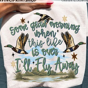 May include: White t-shirt featuring a design with three ducks in flight, a lake scene, and the text "Some glad morning when this life is over I'll Fly Away" in green script. The design includes stars and grass.