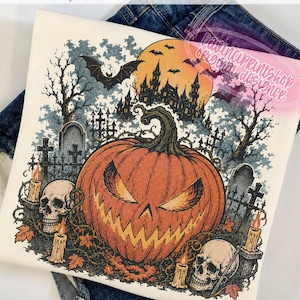 May include: A cream-coloured t-shirt featuring a graphic print of a large orange jack-o'-lantern, two human skulls, lit candles, and a spooky graveyard scene with a castle and a full moon. The shirt is folded over a pair of blue denim jeans.