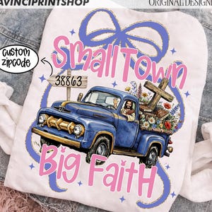 Puede incluir: Camiseta blanca con un diseño de camioneta azul vintage, Jesús conduciendo. La caja de la camioneta está llena de flores, una cruz y libros. El texto "Small Town Big Faith" es en rosa, con un lazo azul y estrellas.