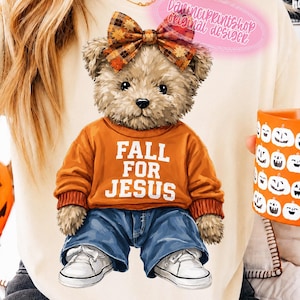 May include: A cream-colored t-shirt featuring a graphic of a teddy bear wearing an orange sweatshirt with the text FALL FOR JESUS, blue jeans, white sneakers, and a plaid bow on its head. The bear is illustrated with brown fur and a soft texture.