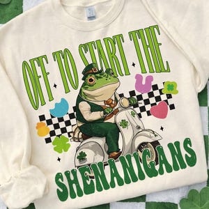 以下が含まれることがあります： クリーム色のスウェットシャツ。聖パトリックの日のデザイン。緑色のカエルがレプラコーンの帽子をかぶり、白いスクーターに乗っています。「OFF TO START THE SHENANIGANS」の文字は緑色で、クローバーなどのお祝いの形が描かれています。