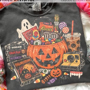 Puede incluir: Una camiseta gris carbón de cuello redondo con un estampado gráfico de un cubo de calabaza lleno de dulces, incluyendo Reese's y M&M's, rodeado por un fantasma, cintas VHS de películas de terror, un café, una cámara y dientes de vampiro.