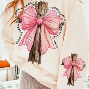 May include: A light beige sweatshirt with a pink bow tied around a wooden cross. The text "She is clothed with strength and dignity and she laughs without fear of the future Proverbs 31:25" is printed around the cross.