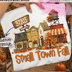 May include: A white t-shirt featuring a colorful graphic of a small town street scene during autumn. The design includes houses, a water tower, pumpkins, fall foliage, a wooden sign reading 37203 Nashville TN, and the text Small Town Fall.