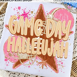 May include: A white t-shirt featuring a graphic print of a large brown star, two pink football helmets, and pink star accents. The text GAME DAY HALLELUJAH is printed in large, light tan letters across the center of the design.